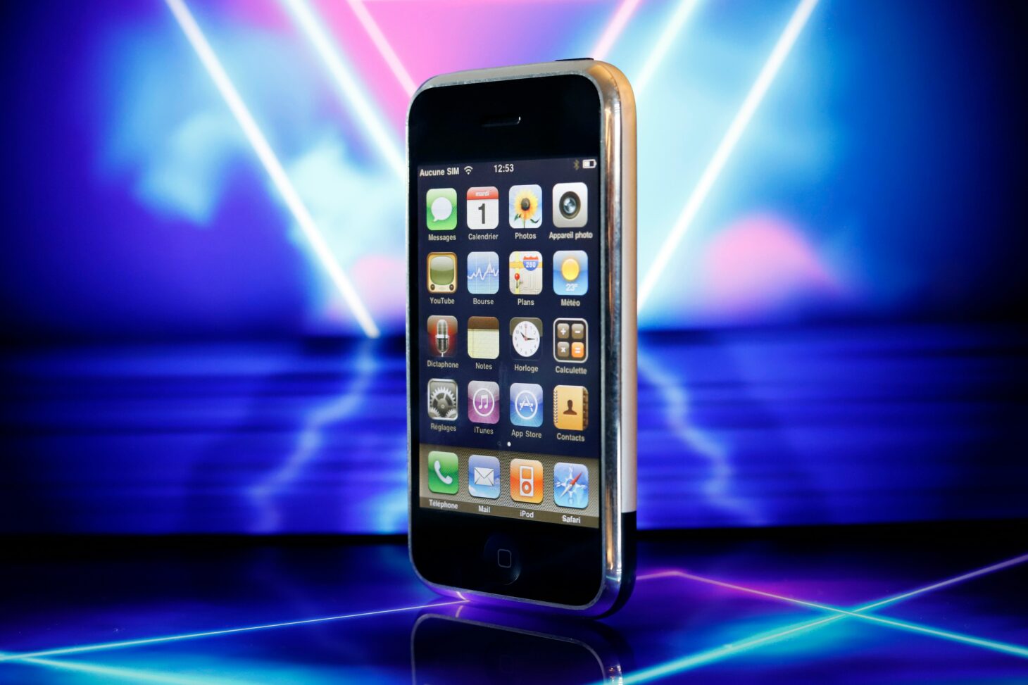 ネオンの背景に立てた初代iPhone。クロムのフレームとホームボタン、iPhone OSのフランス語ホーム画面が見える
