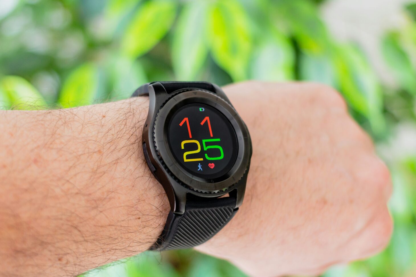 緑の葉を背景に手首に着けた黒い丸型スマートウォッチ。カラフルな数字の文字盤を表示している