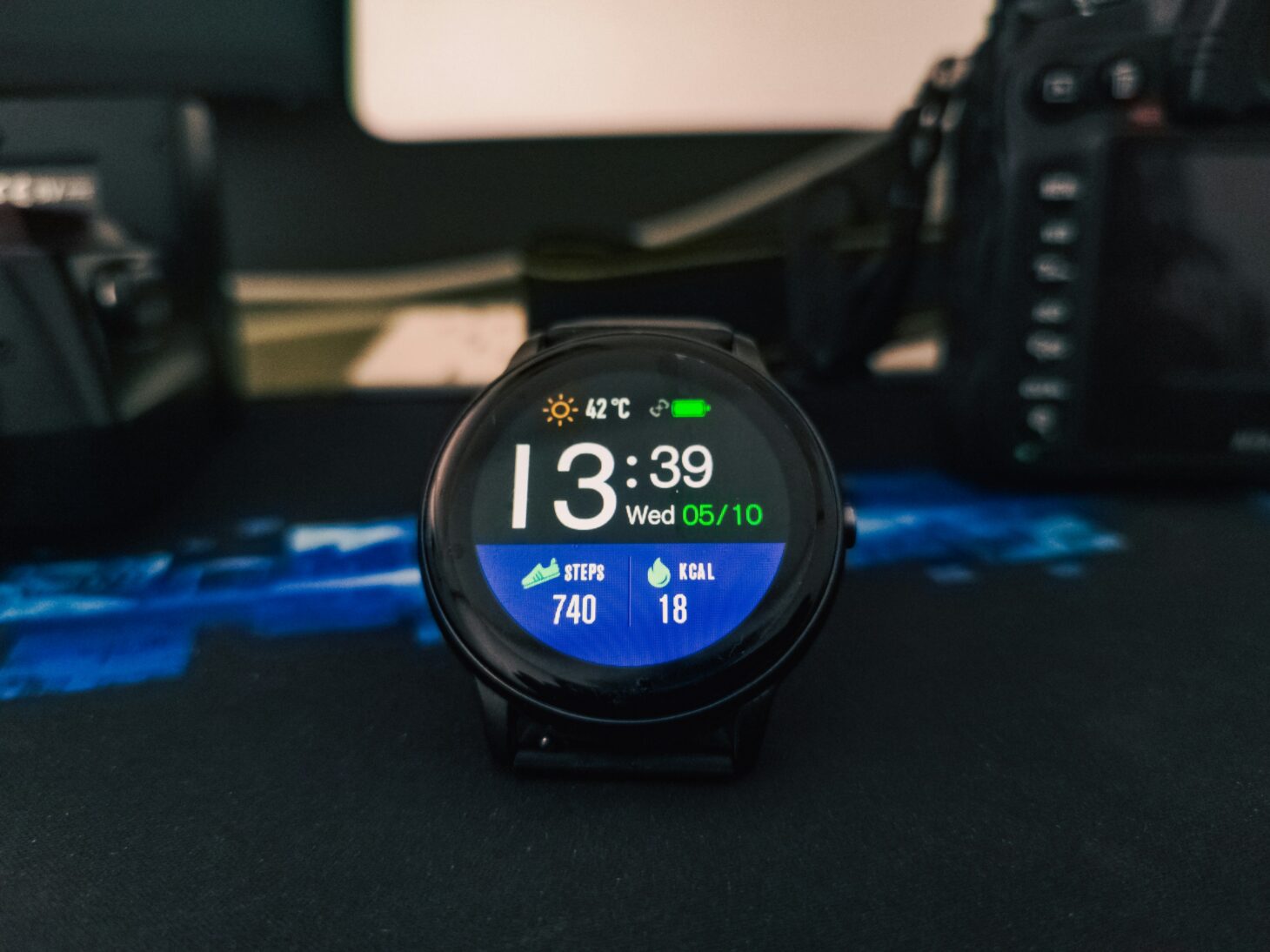 机に置かれた黒い丸型スマートウォッチ。天気・時刻・歩数740・18kcalが表示されている