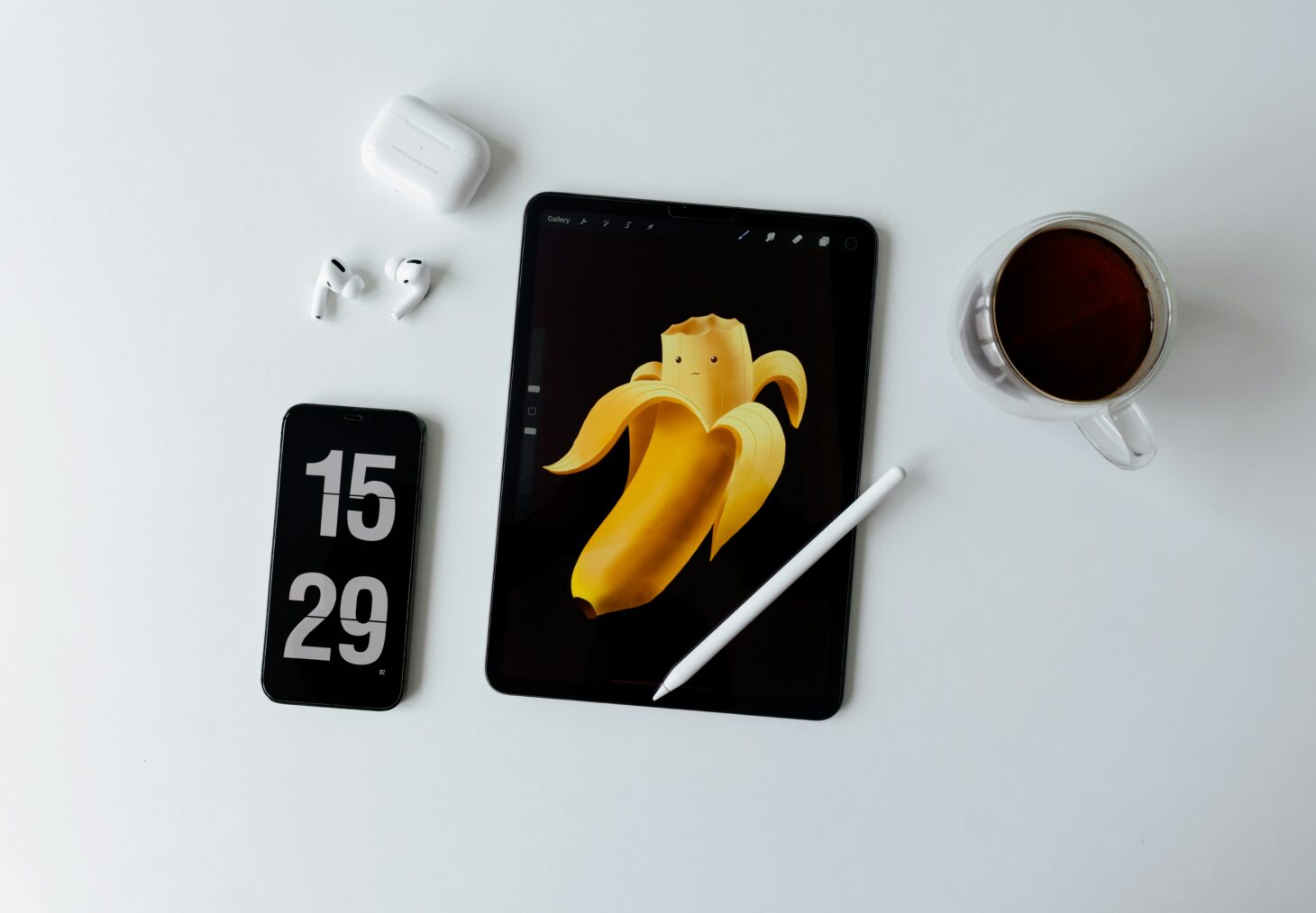 白い机に並んだiPad(Apple Pencil・バナーのイラスト表示)、フリップ時計表示のiPhone、AirPods Pro、コーヒーカップ。Apple Watchは写っていない。