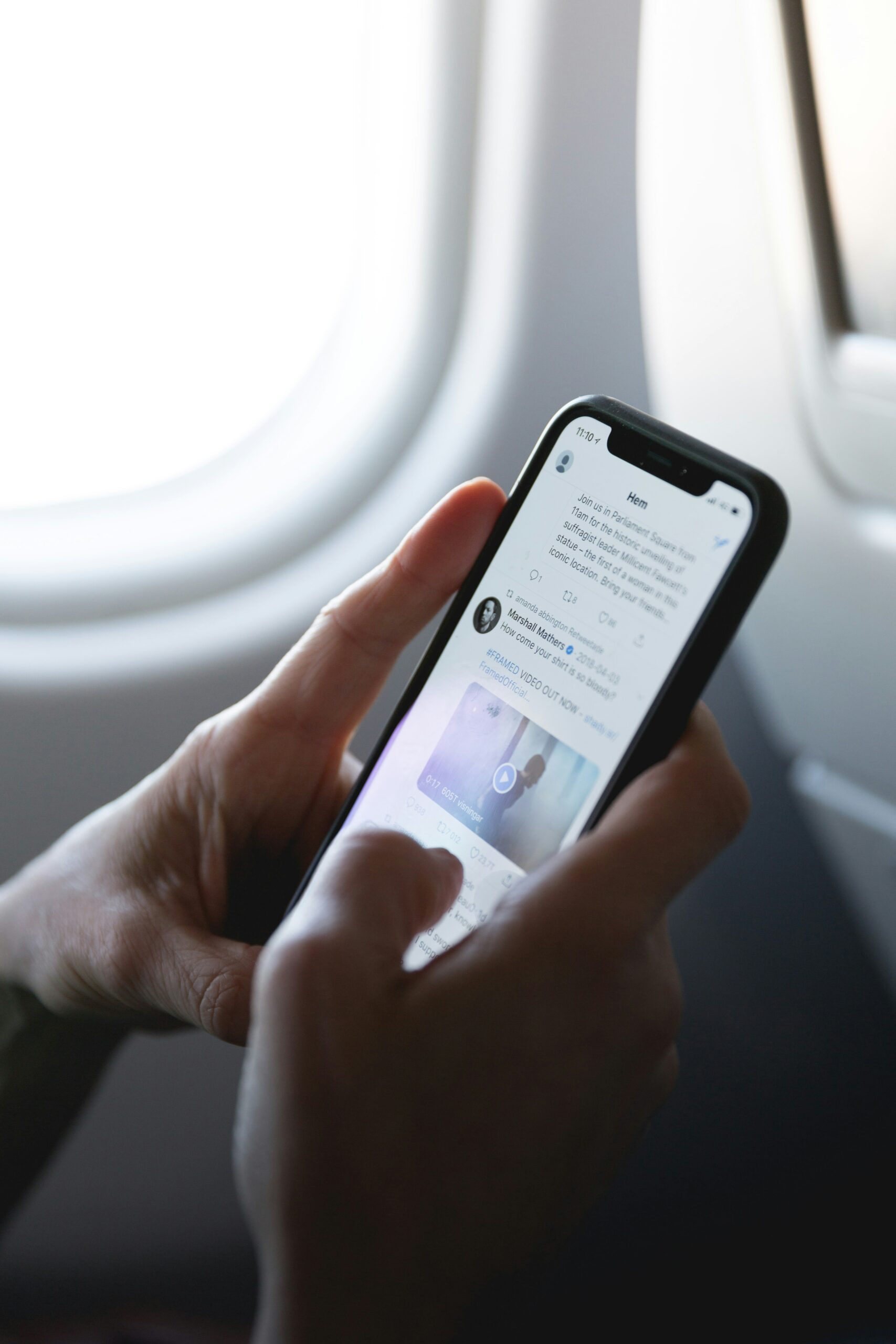飛行機の窓際の席でノッチ付きiPhoneのSNSを両手で操作している様子