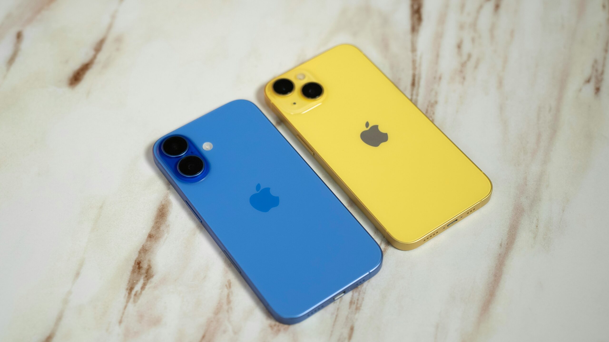 大理石調の面に並べたブルーのiPhone 16とイエローのiPhone 14系の背面