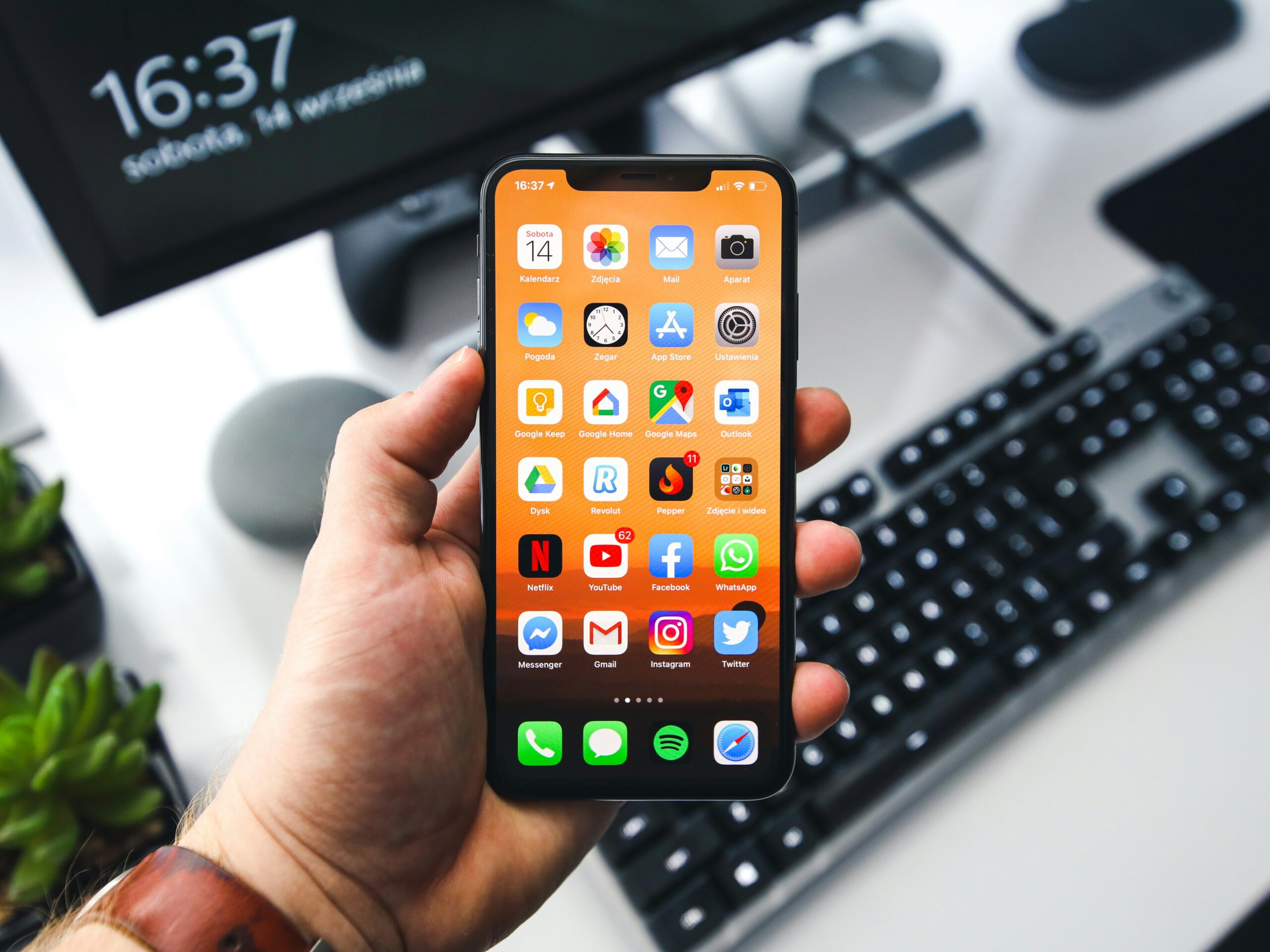 デスクのキーボードの前でノッチ付きiPhoneを片手で持ち、オレンジ色の壁紙のホーム画面を表示している