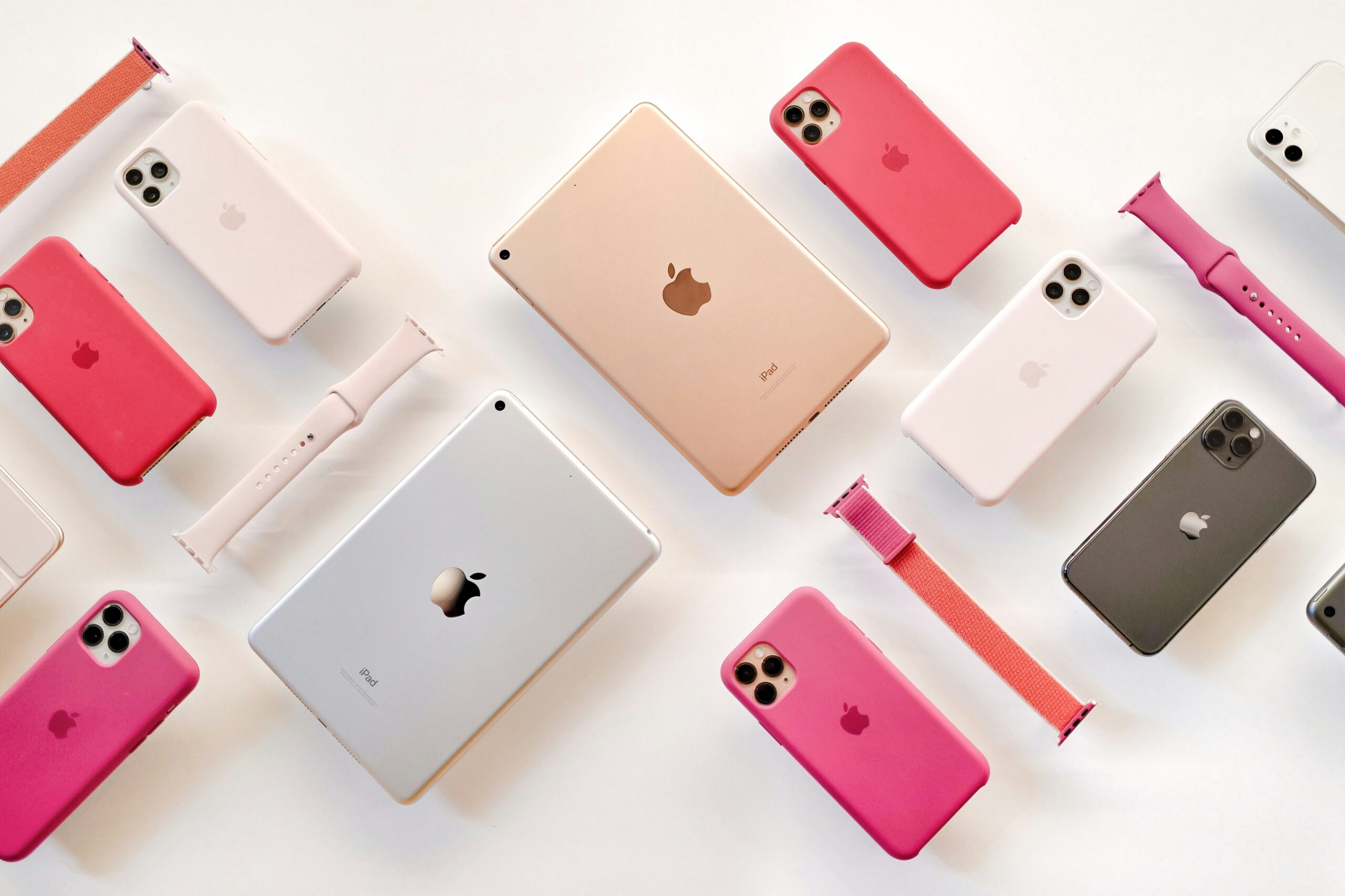白い背景にピンク系の純正シリコンケースを付けたiPhone 11 Pro系、ゴールドとシルバーのiPad、Apple Watchのバンドを並べた俯瞰写真