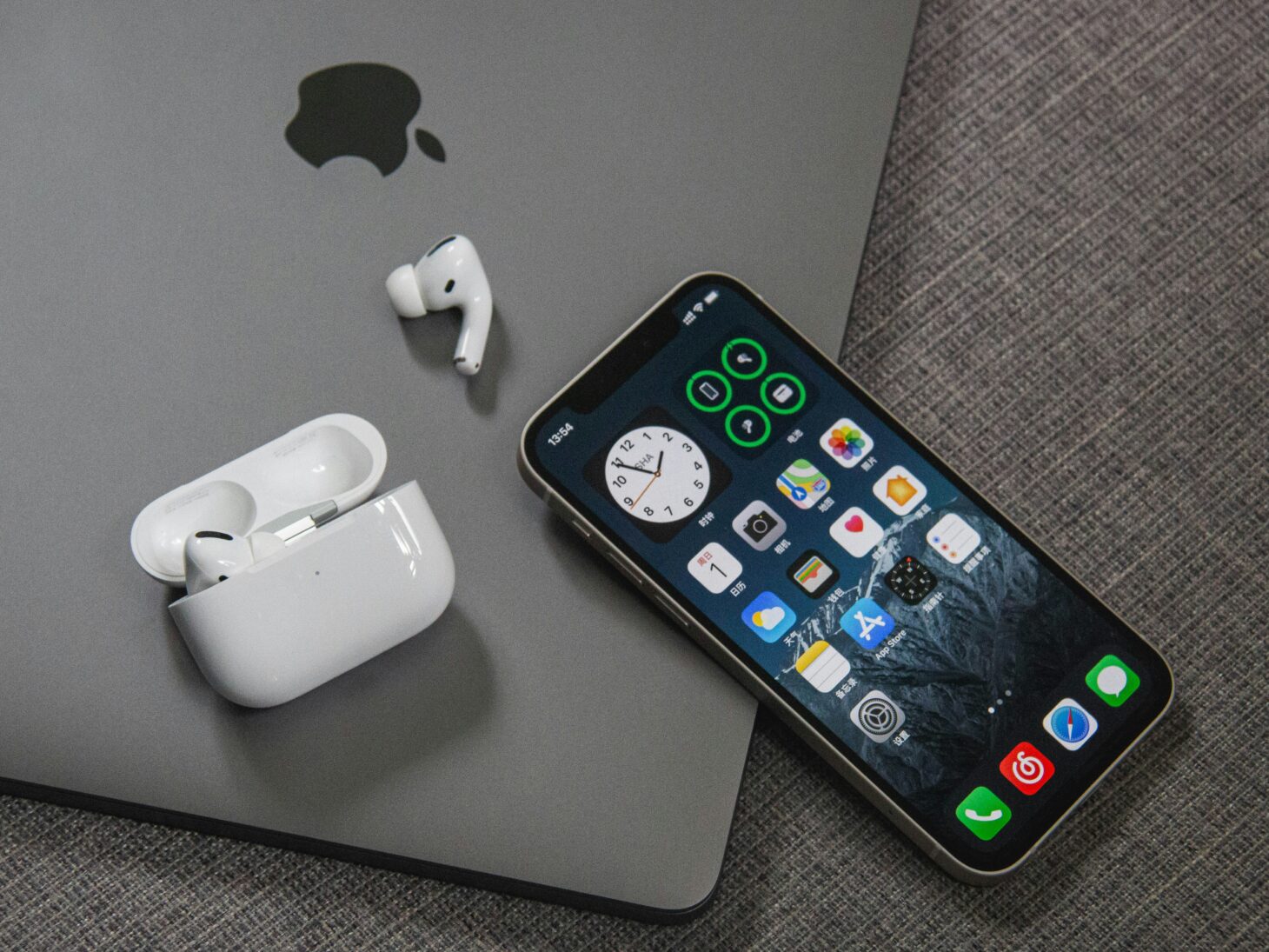 MacBookの上に置いたノッチ付きiPhoneのホーム画面と、AirPods Proのケースとイヤホン