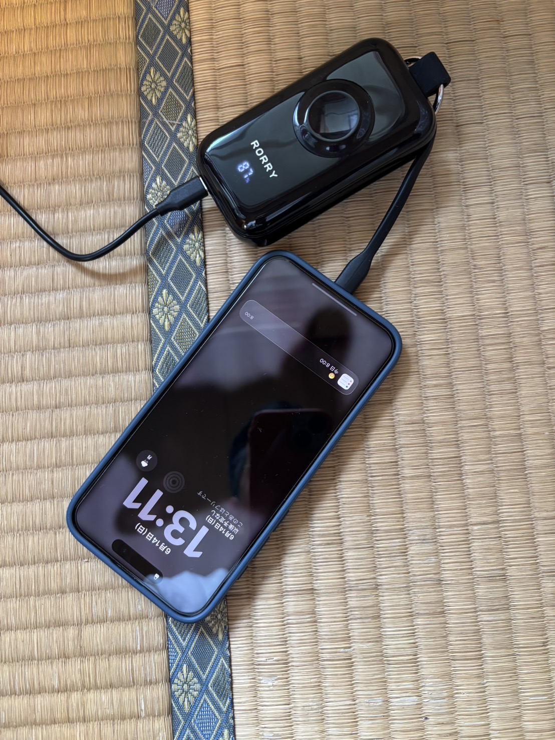 畳の部屋でRORRY CharmGo D4-10000がコンセントに繋がれつつiPhoneにもケーブル給電しているパススルー充電のシーン。本体残量は81%