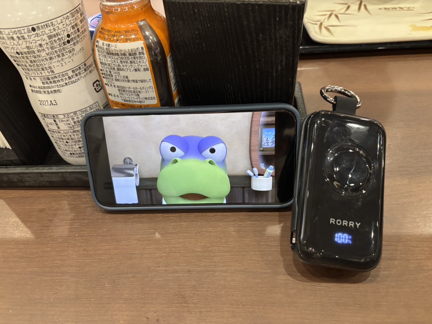 うどん屋のテーブルでiPhoneにアニメを表示しながらRORRY CharmGo D4-10000で充電している様子。バッテリー残量は100%表示