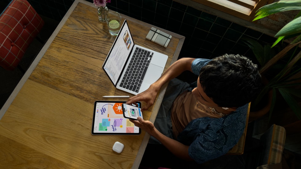 iPhoneを手に持ち、テーブルでMacBook Proを使って作業している人物を上から見た様子。周りにiPad、Apple Pencil、AirPodsが置かれている