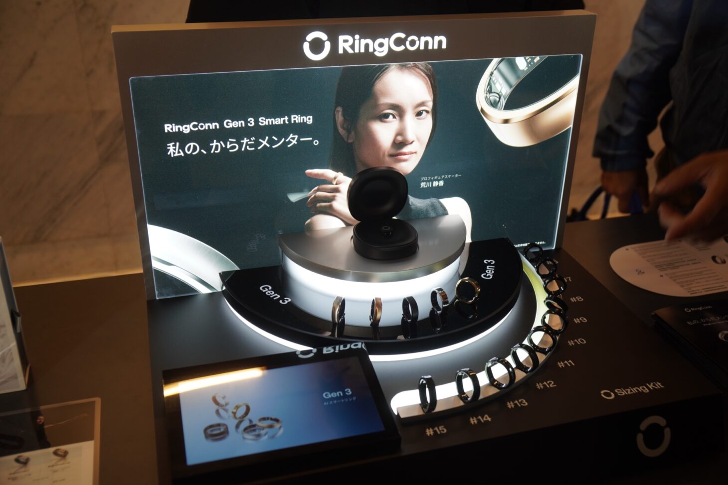 RingConn Gen 3の製品展示ブース全景。「私の、からだメンター」のポスター、5色のリング実機、サイジングキット(リング15サイズ並び)、AIスマートリング Gen 3 のディスプレイ