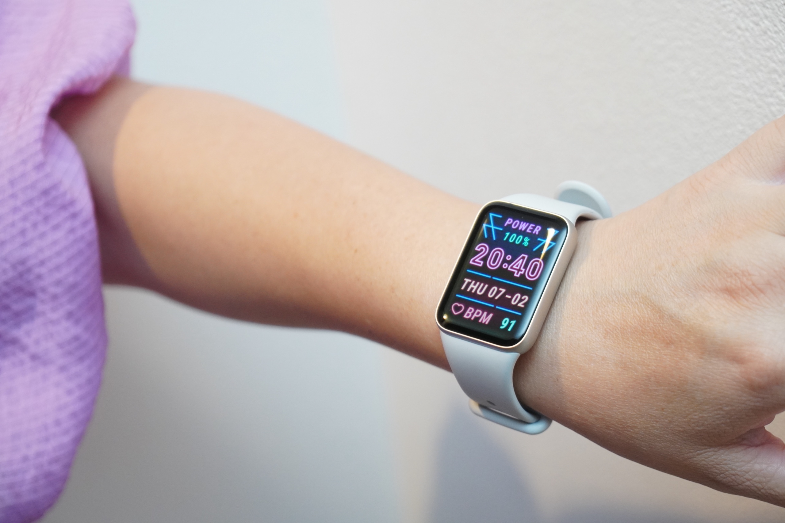 シルバーの Xiaomi Smart Band 10 Pro にピンク文字のウォッチフェイスを表示し、ラベンダーの服に合わせた様子