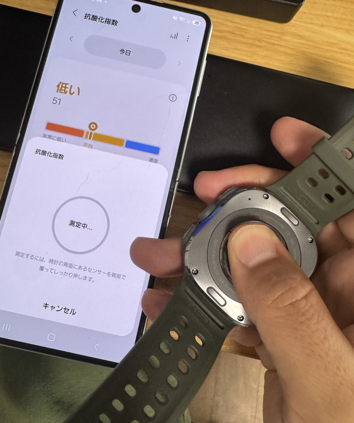 Galaxy Watch Ultra2の背面センサーを親指で押して抗酸化指数を測定しているところ。スマートフォンには「測定中…」と、時計の背面にあるセンサーを親指で覆ってしっかり押す旨の案内が出ている。
