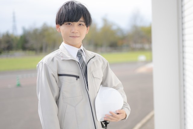 ヘルメットを手に持ち笑顔で立つ作業服姿の若い男性