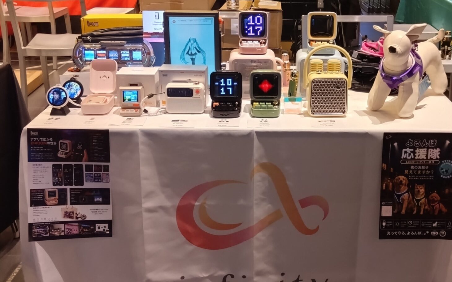 ガジェット展示会2026 秋のinfinityブース。テーブルにDivoomのピクセルディスプレイ搭載スピーカーや時計が並び、右側にLEDドッグハーネスを着けた犬のマネキンが置かれている