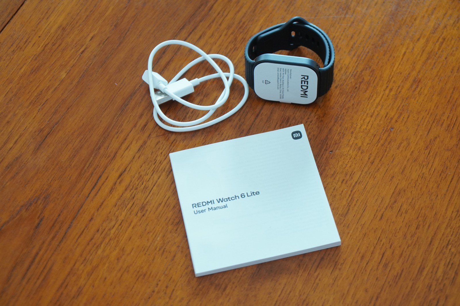 REDMI Watch 6 Liteの同梱品。ストラップを付けた本体、白い磁気充電ケーブル、正方形の取扱説明書が木のテーブルに並んでいる