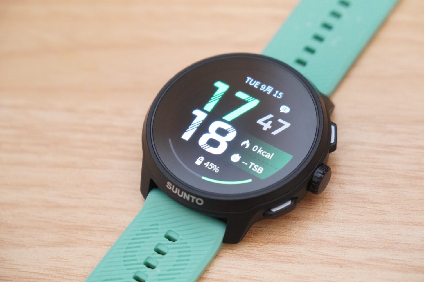 Suunto Run 2の本体を木のテーブルに置いた状態。黒いケースにミントグリーンのシリコンストラップが対角に伸び、画面に時刻とバッテリー残量が表示されている