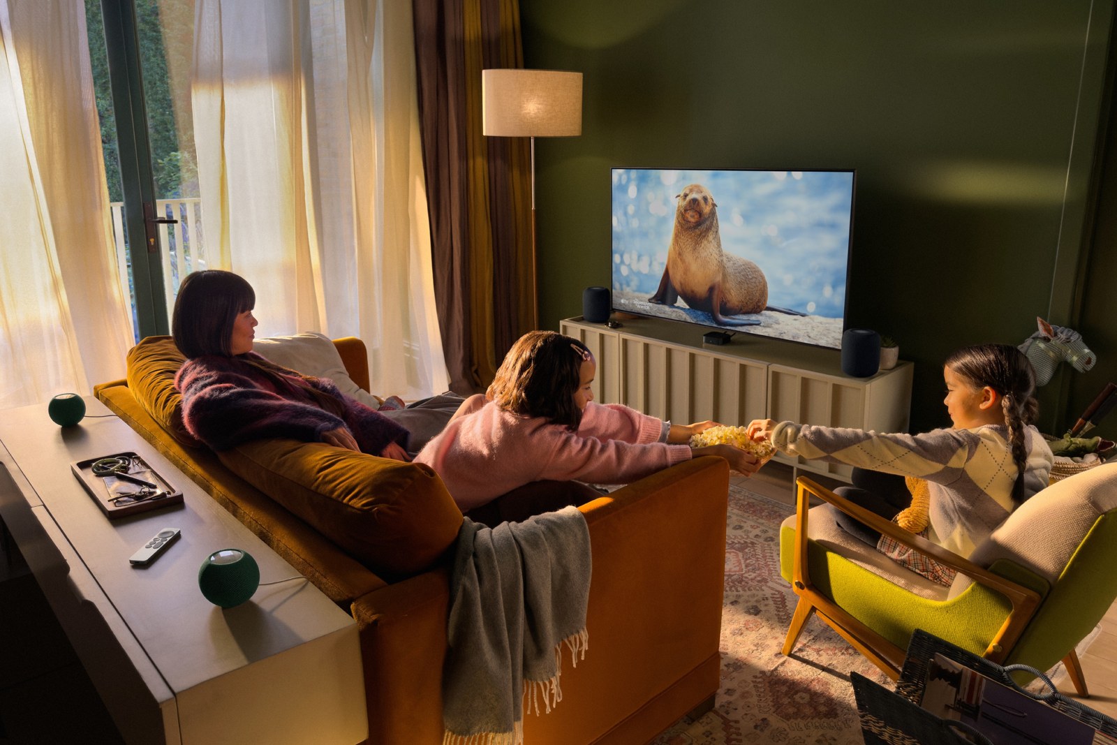 リビングのテレビの左右と後ろにHomePodを置き、家族がソファでApple TV 4Kの映像を観ている様子