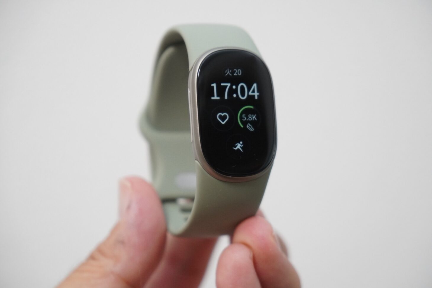 手に持ったGoogle Fitbit Edgeの正面。金属のケースに縦長の画面があり、17:04の時刻と歩数が表示されている。バンドは淡いグリーン