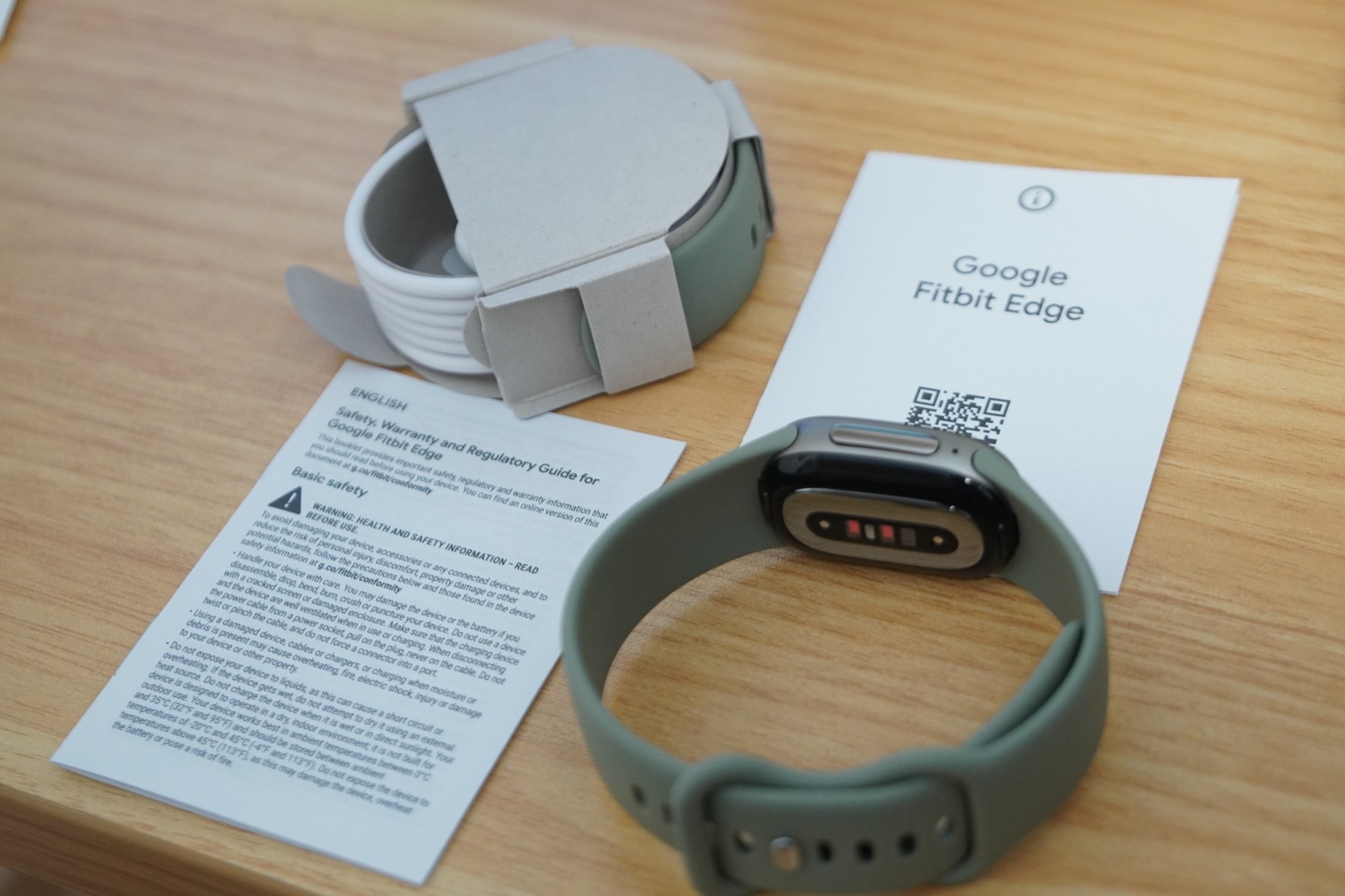 机に並べたGoogle Fitbit Edgeの同梱物。紙で巻かれた充電ケーブル、安全と保証のガイド、Google Fitbit Edgeと書かれたカード、裏面のセンサーが見える本体