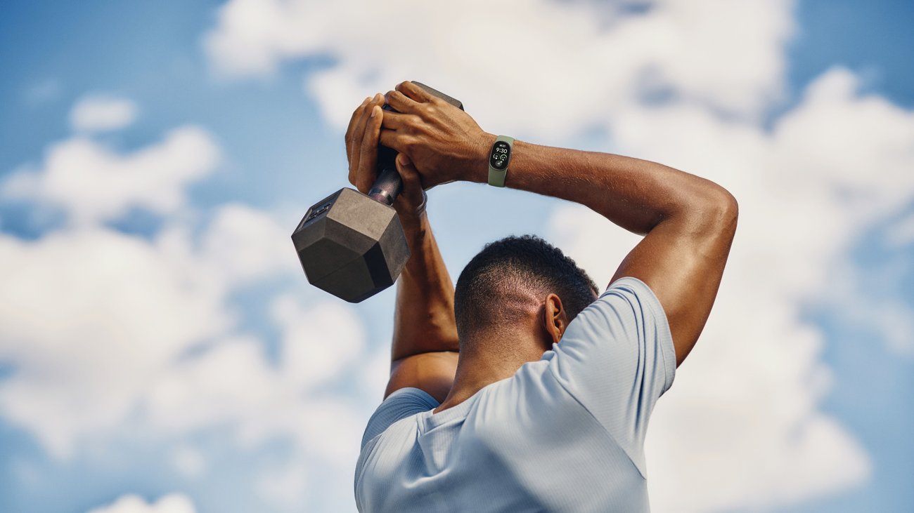ダンベルを頭の後ろに持ち上げる男性の手首に、オリーブ色のバンドのGoogle Fitbit Edgeが着いている