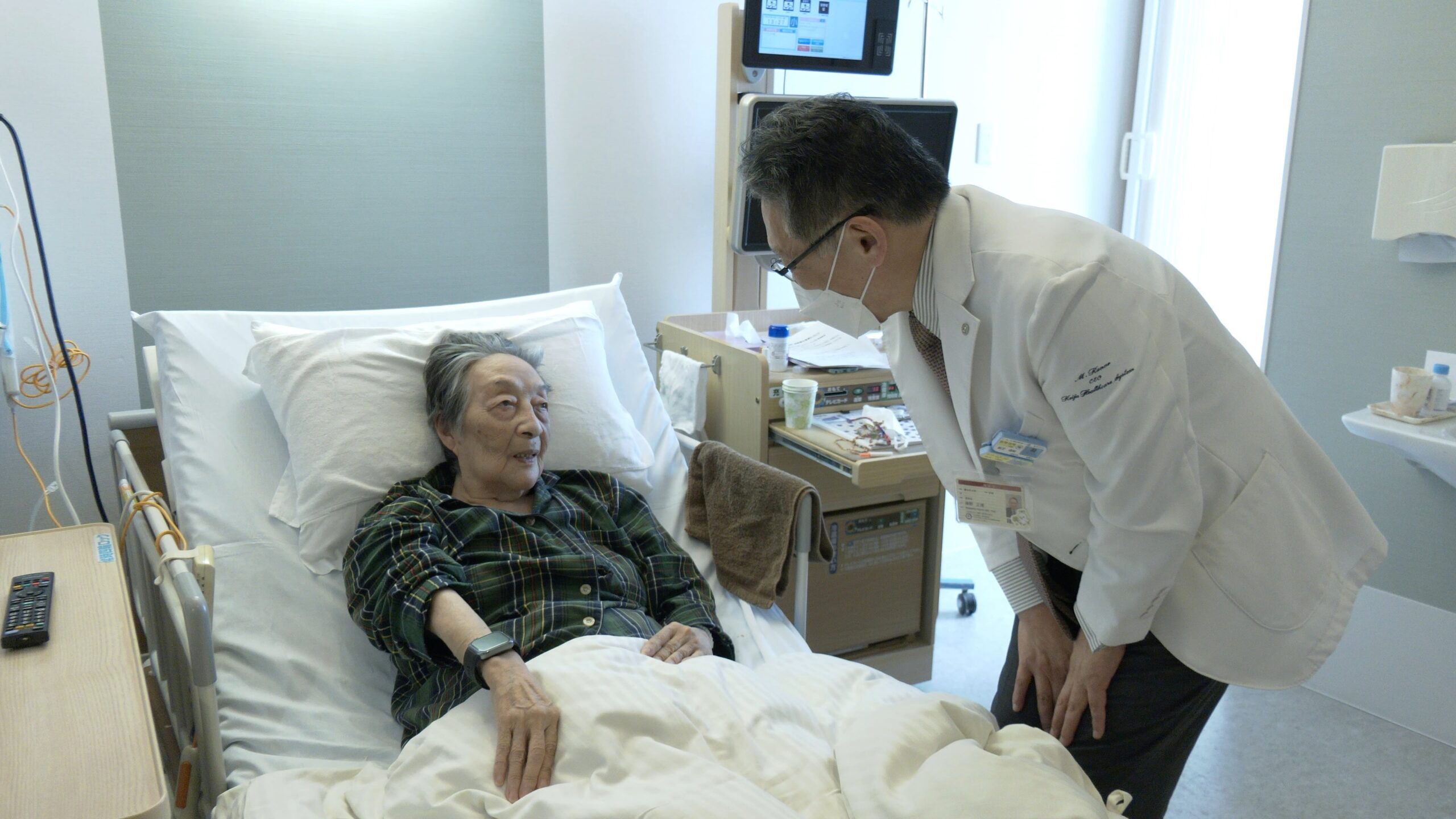 恵寿総合病院の病室で、腕にスマートウォッチを着けてベッドに横になる入院患者に医師が話しかけている様子