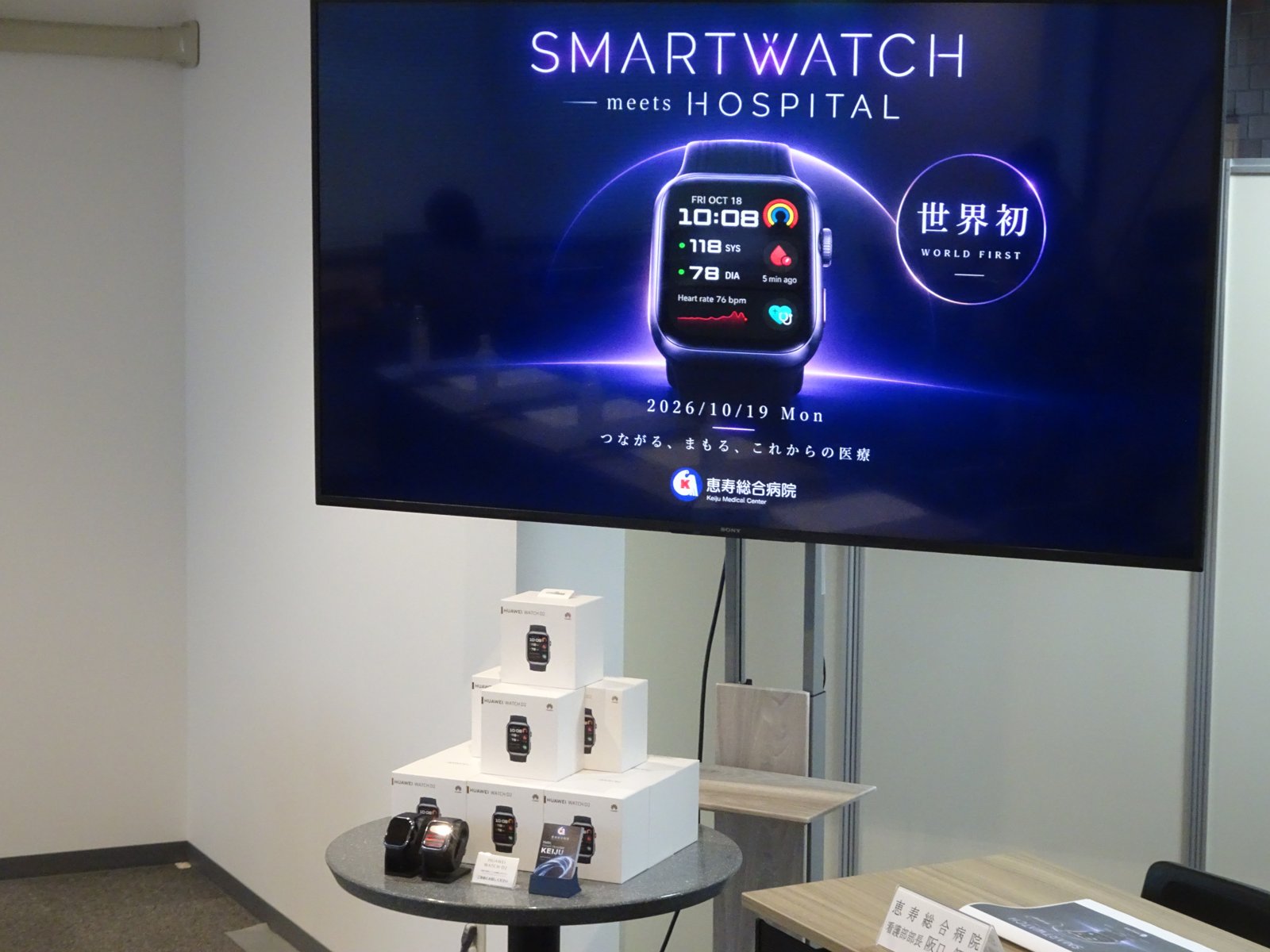 会見場の画面に映した「SMARTWATCH meets HOSPITAL」「世界初」のスライドと、台に並べたHUAWEI WATCH D2の箱と本体