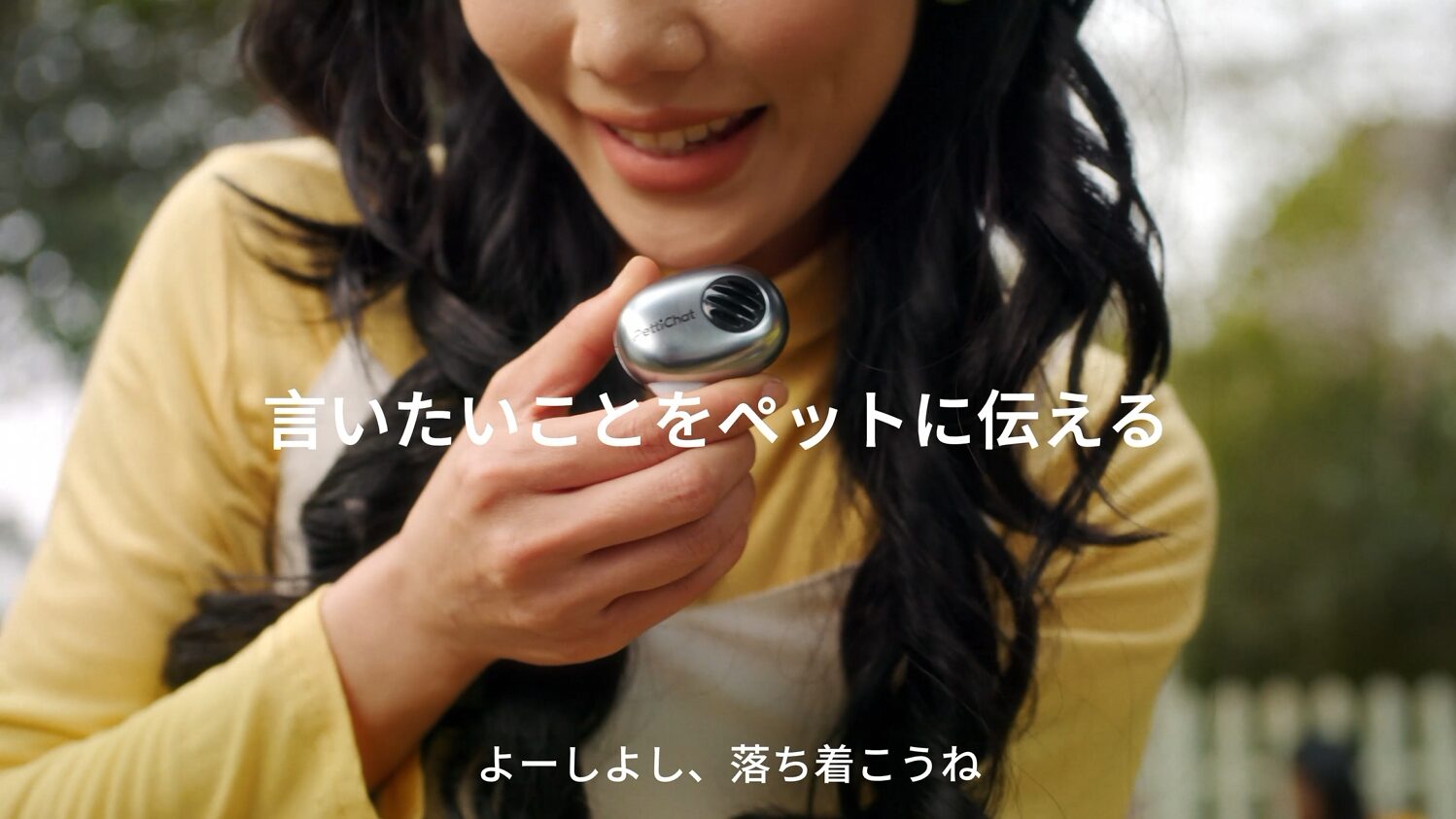 シルバーのPettiChat本体を手に持って話しかける女性。画面に「言いたいことをペットに伝える」「よーしよし、落ち着こうね」の文字