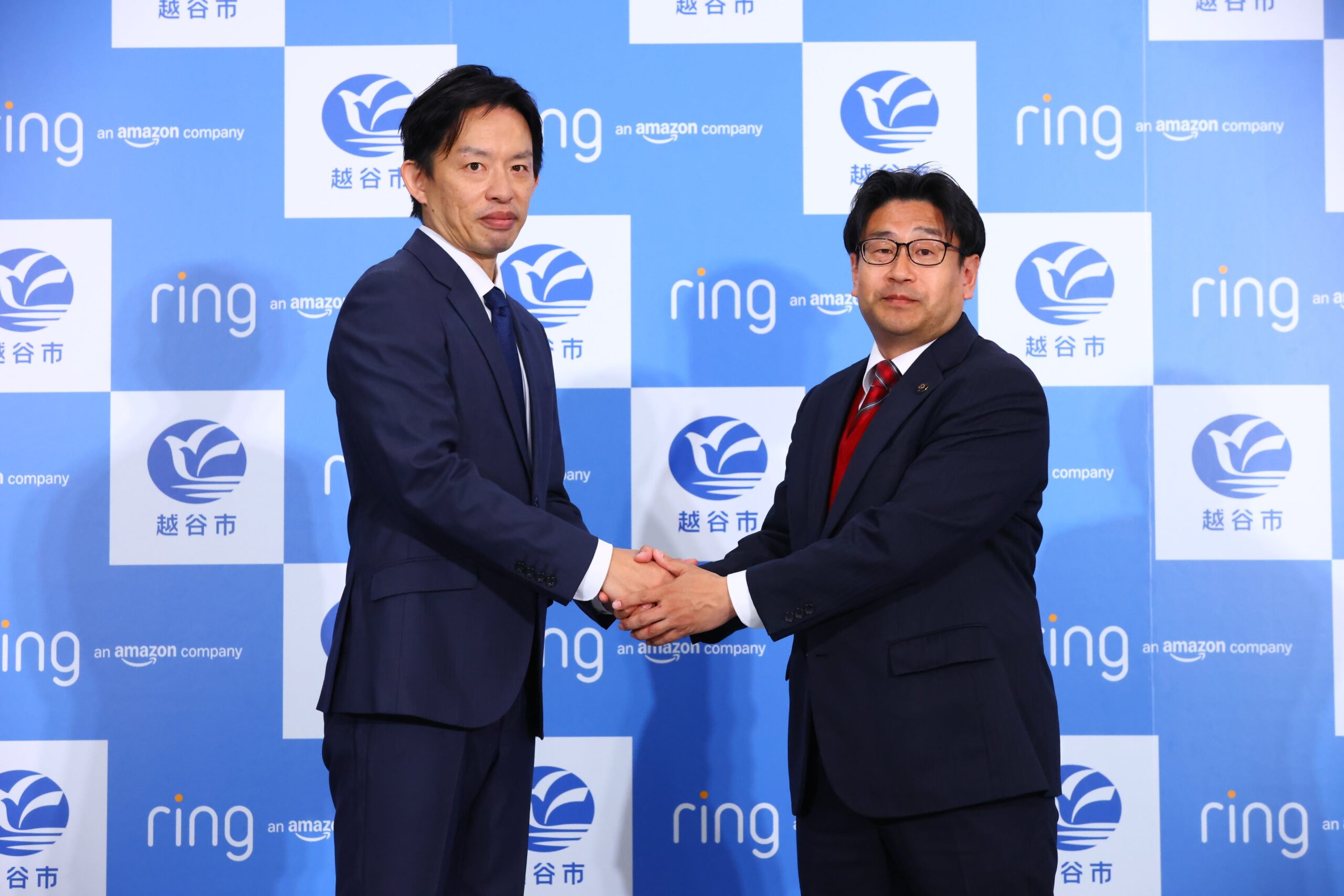 アマゾンジャパンRing事業部の瀬口雄介さんと越谷市長の福田晃さんが、Ringと越谷市のロゴの前で握手している様子