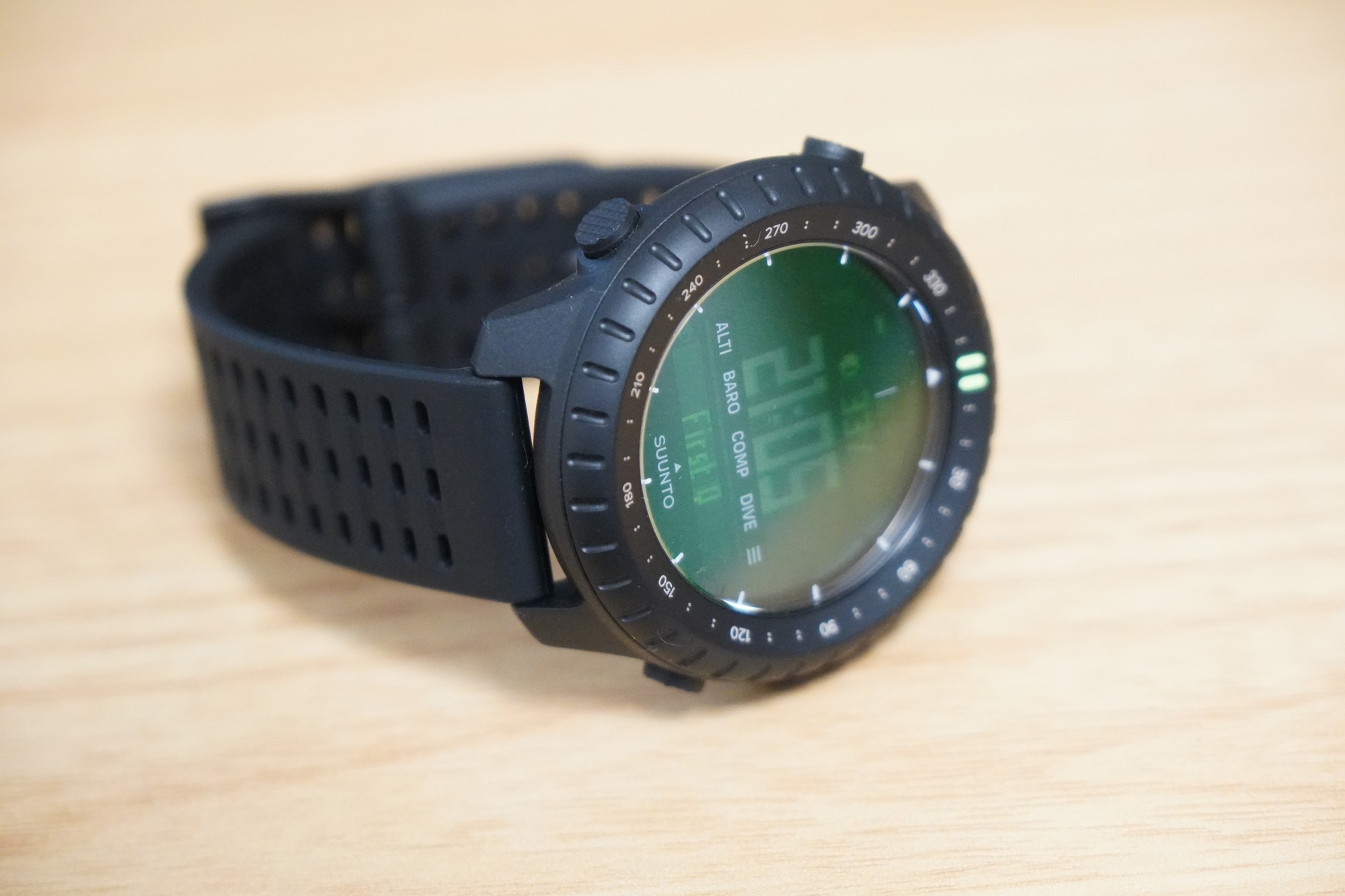 机の上に置いたSuunto Core 2。緑のバックライトが点いた画面と、左側のボタン、穴の並んだシリコンストラップが写っている