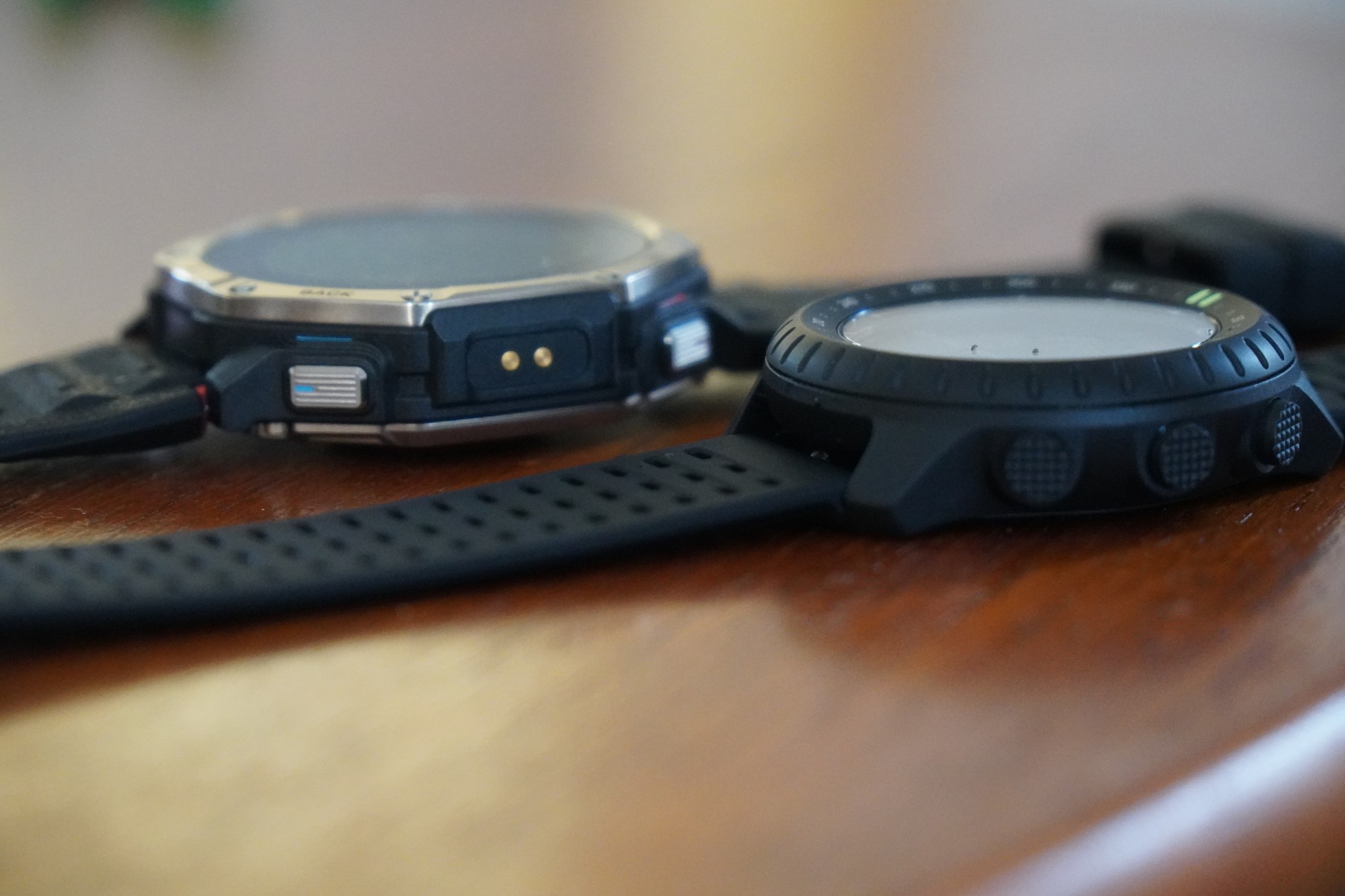 机の上に横から並べたAmazfit T-Rex Dual Solar(左)とSuunto Core 2(右)。T-Rexの側面には金色の2つの接点、Core 2の側面には3つのボタンが見える
