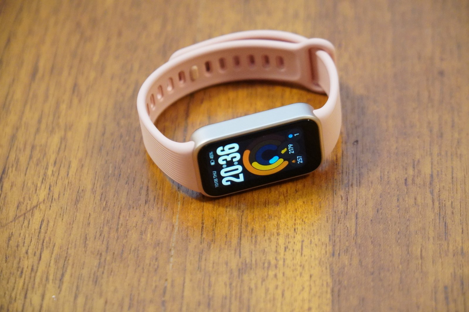 木の机に置いたXiaomi Smart Band 11 Activeのポメロピンク。縦長の画面に20時36分の時刻と3色のリングが表示されている